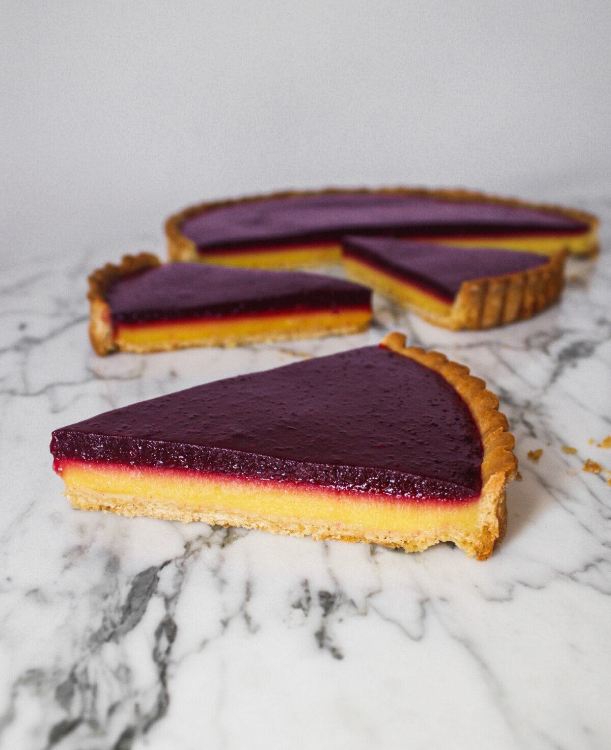Blueberry lemon tart with yellow and purple layers, set on a gray plate on top of a marble surface.