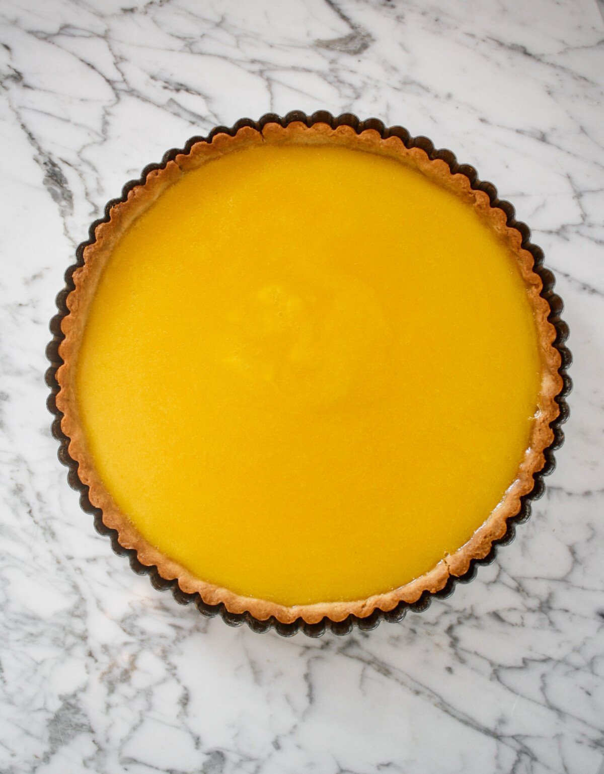 Tart pan lined with baked tart crust and filled with lemon curd set on top of a marble surface.