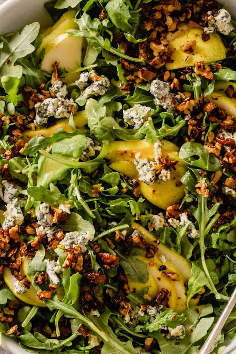 arugula, pears and crumbled blue cheese in a large white bowl with two serving spoons set in it