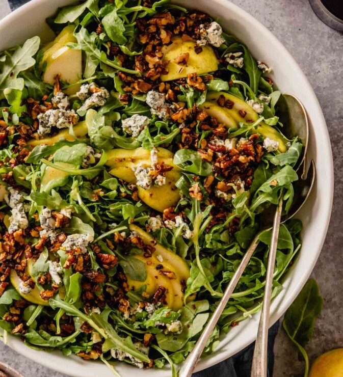 arugula, pears and crumbled blue cheese in a large white bowl with two serving spoons set in it