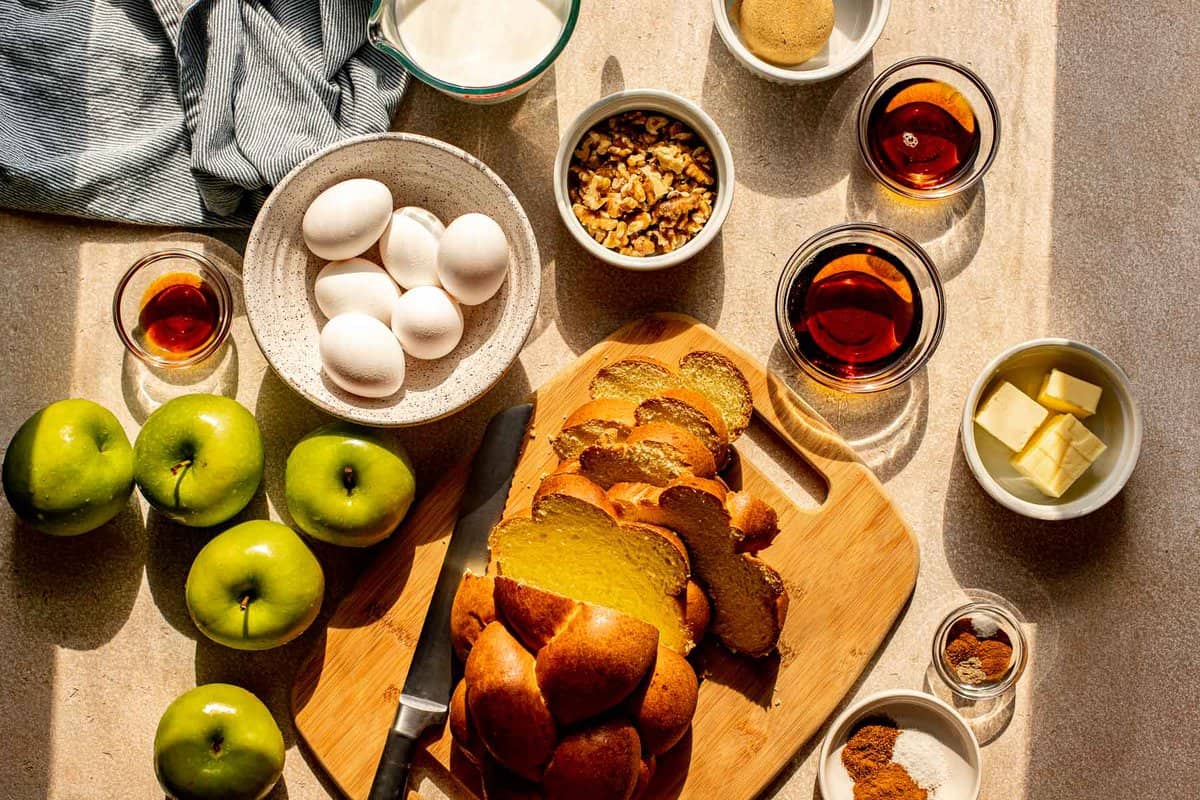 sliced challah bread, apples, eggs, sugar, spices, and maple syrup set out on a counter