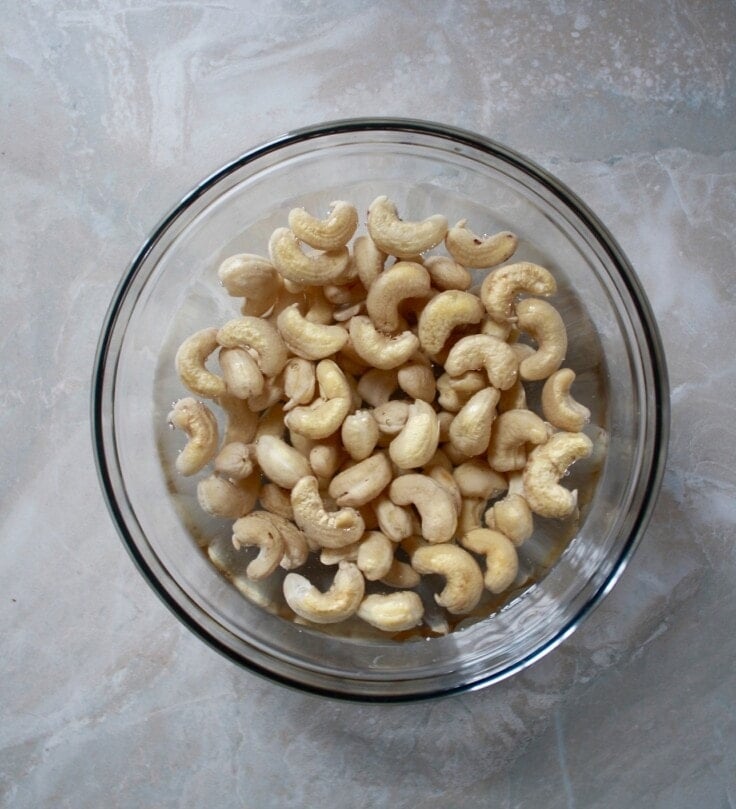 Glass jar filled with water and whole cashews set on a marble surface.