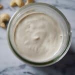 Glass jar filled with smooth cashew cream set on a marble surface with whole cashews set around.