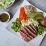 Photograph wasabi crusted tuna set on a white place with brown rice, carrots, cabbage, cucumbers and pickled ginger.