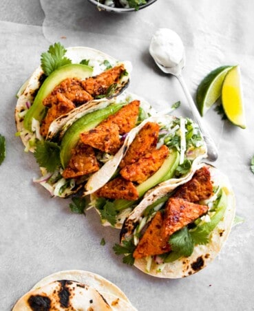 Overhead photo of vegan tempeh tacos arranged in a line across white paper with limes and a creamy sauce off to the side