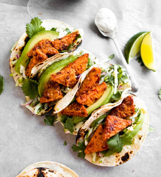Overhead photo of vegan tempeh tacos arranged in a line across white paper with limes and a creamy sauce off to the side