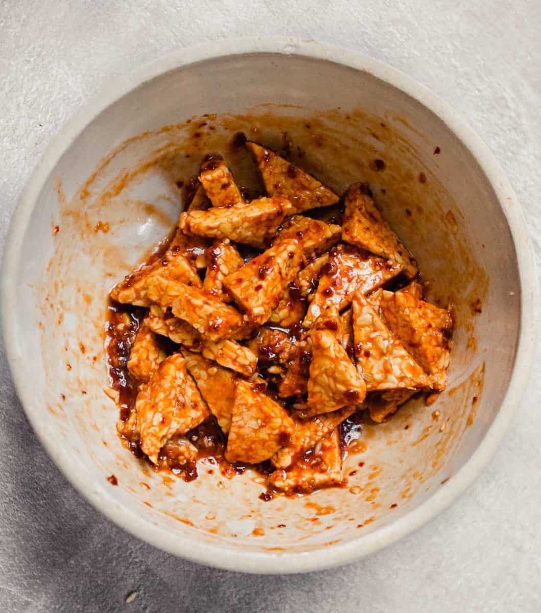 Photograph of tempeh marinated in a red sauce in a cream-colored bowl