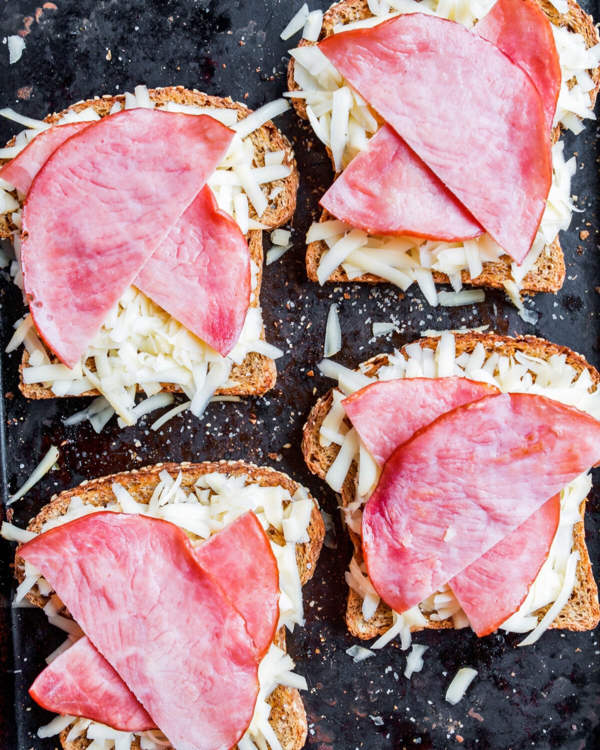 Toasted bread with ham and shredded cheese on black baking sheet