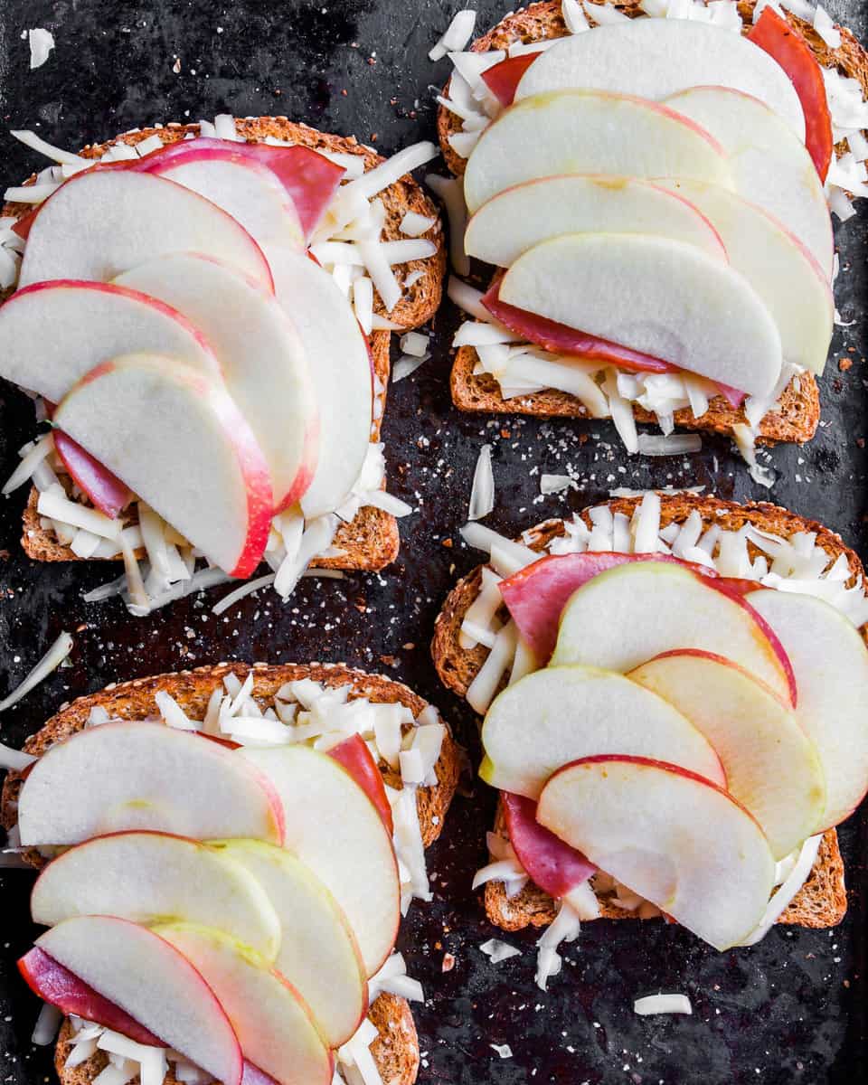 Toasted bread with ham, shredded cheese, and apple on black baking sheet