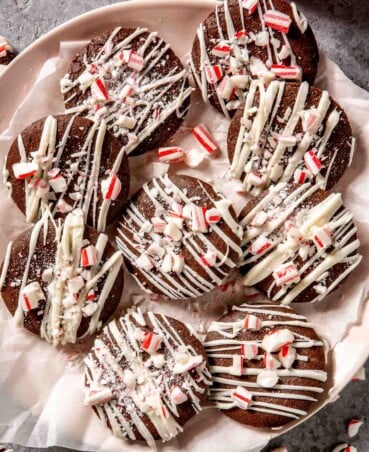 chocolate cookies topped with white chocolate drizzle and crushed peppermint candies set on a parchment-lined plate