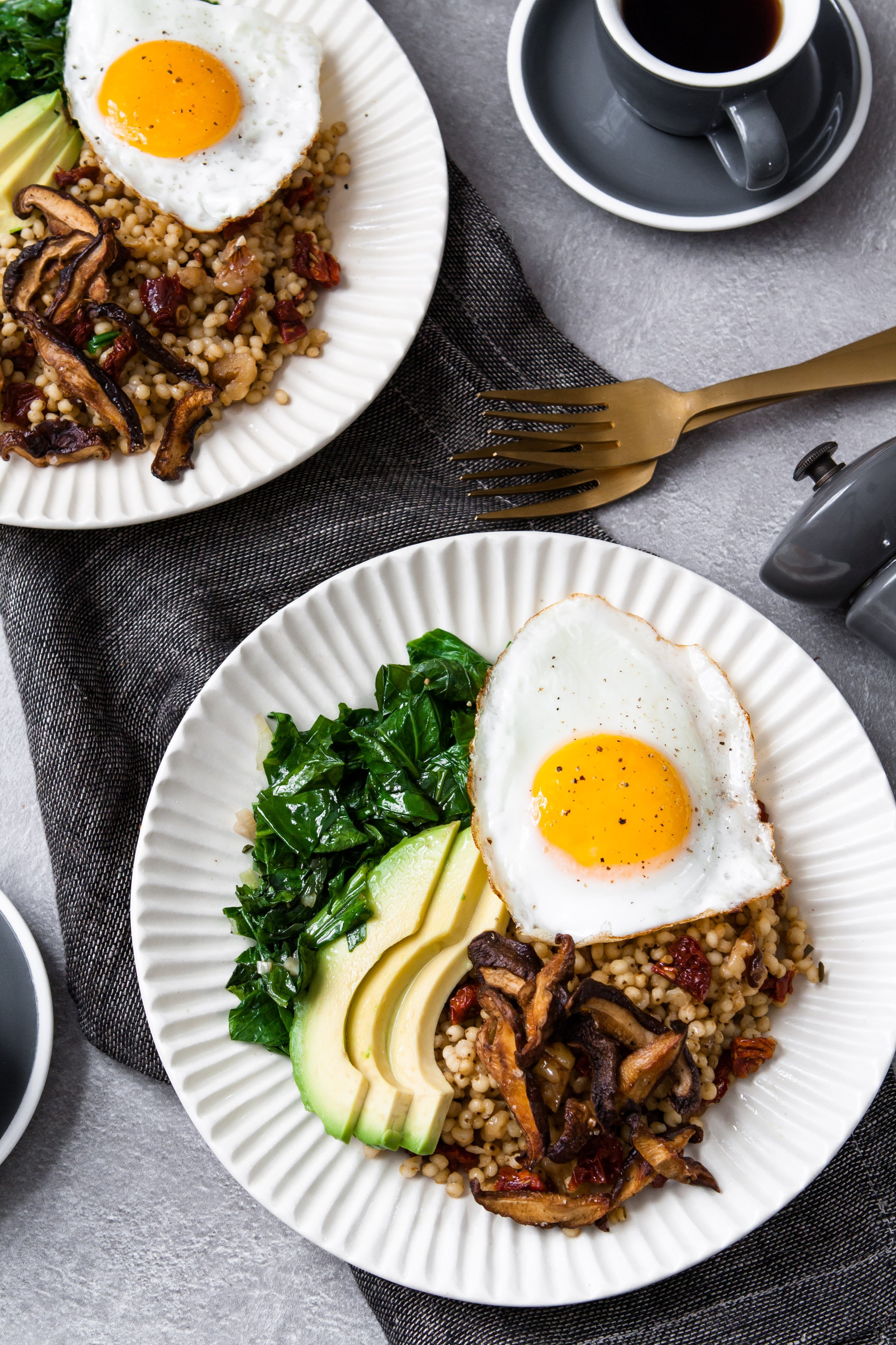 two white bowls filled with grains, sautéed greens, sliced avocado, and topped with a sunny side up egg