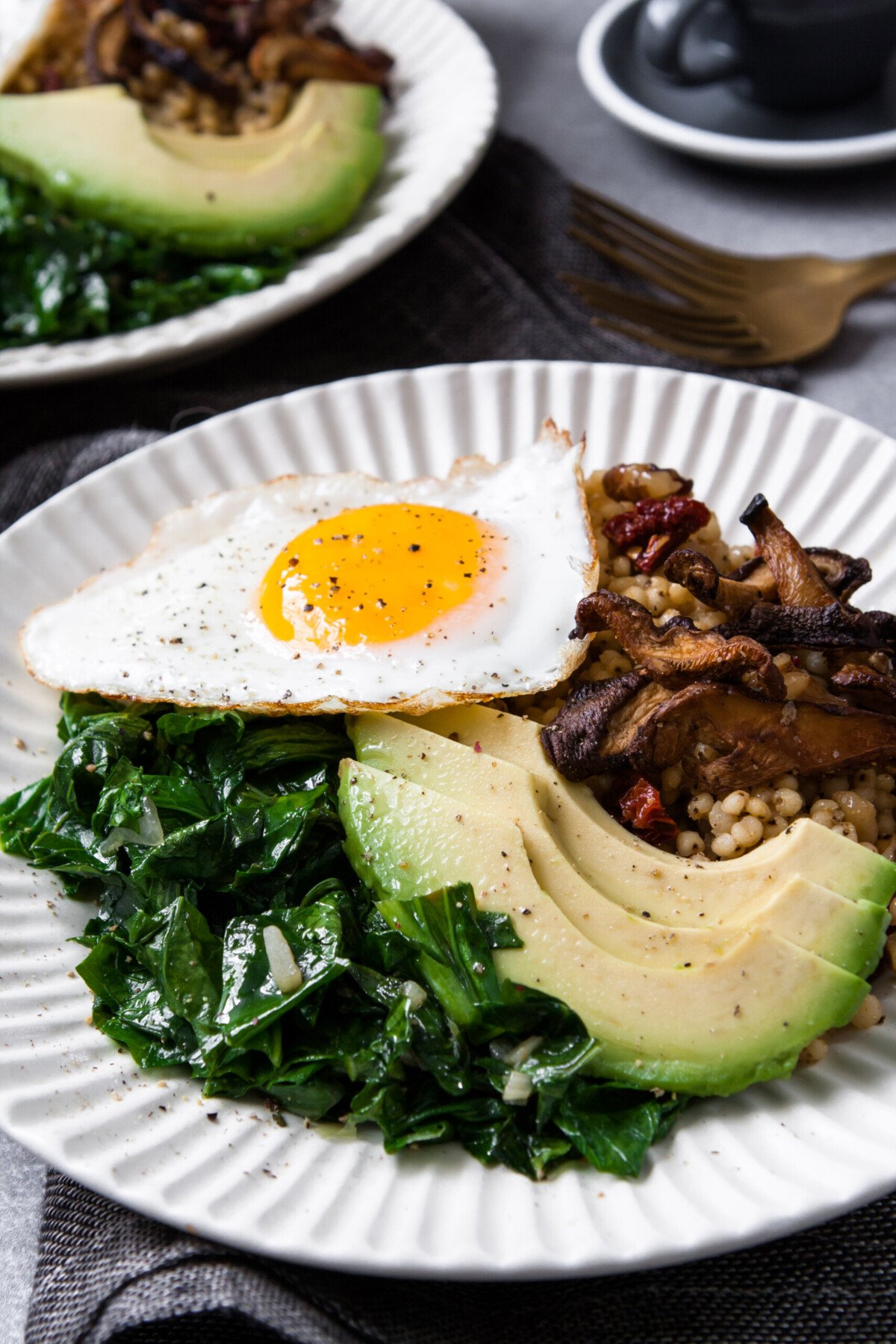  two white bowls filled with grains, sautéed greens, sliced avocado, and topped with a sunny side up egg