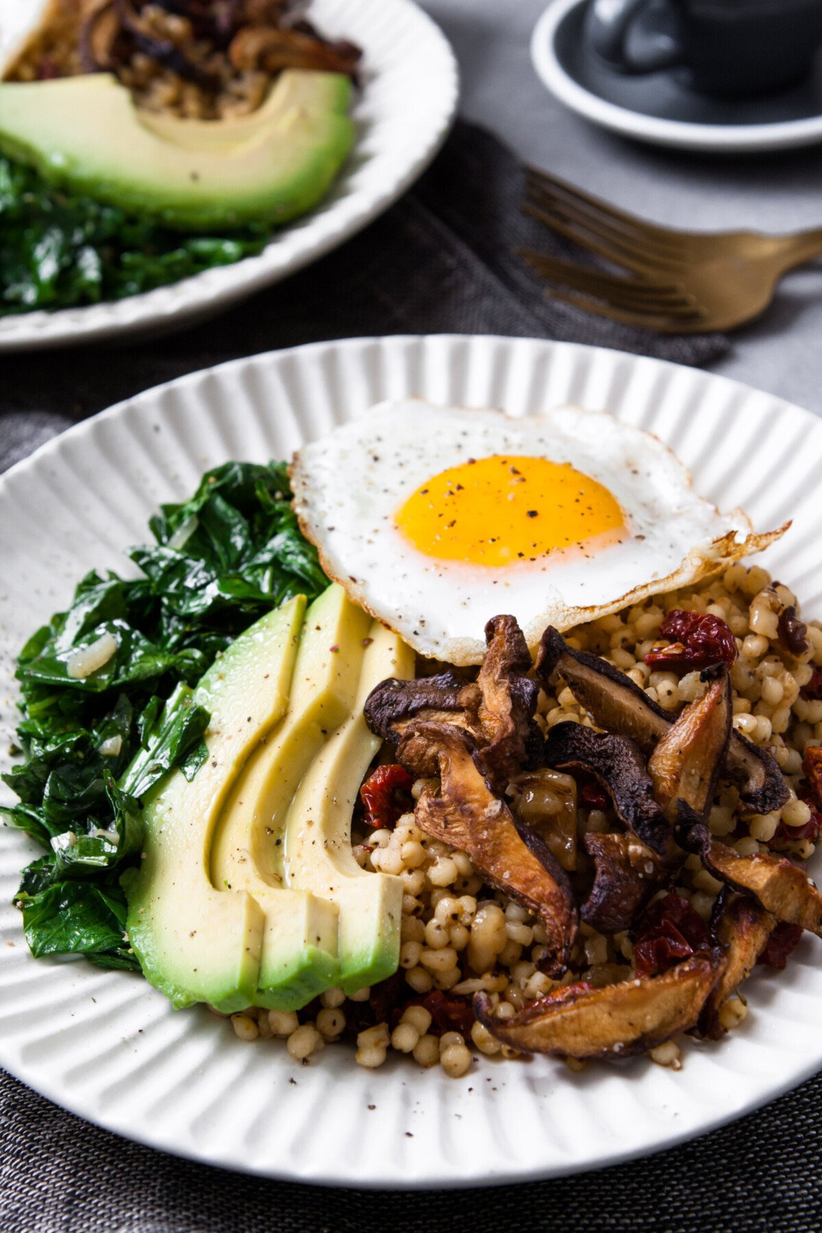 two white bowls filled with grains, sautéed greens, sliced avocado, and topped with a sunny side up egg