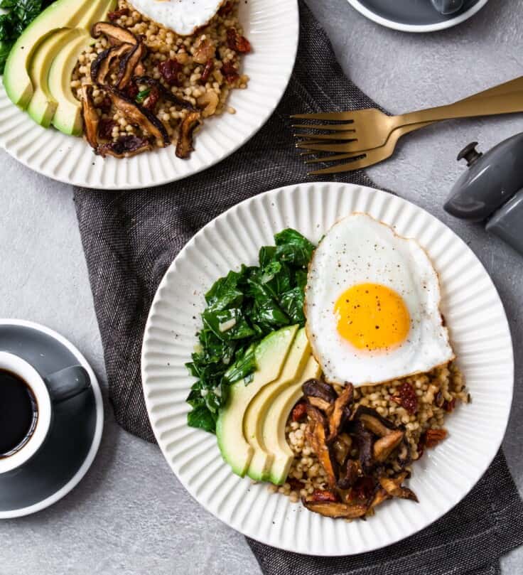 two white bowls with whole grains, sliced avocado, sautéed greens, topped with an egg