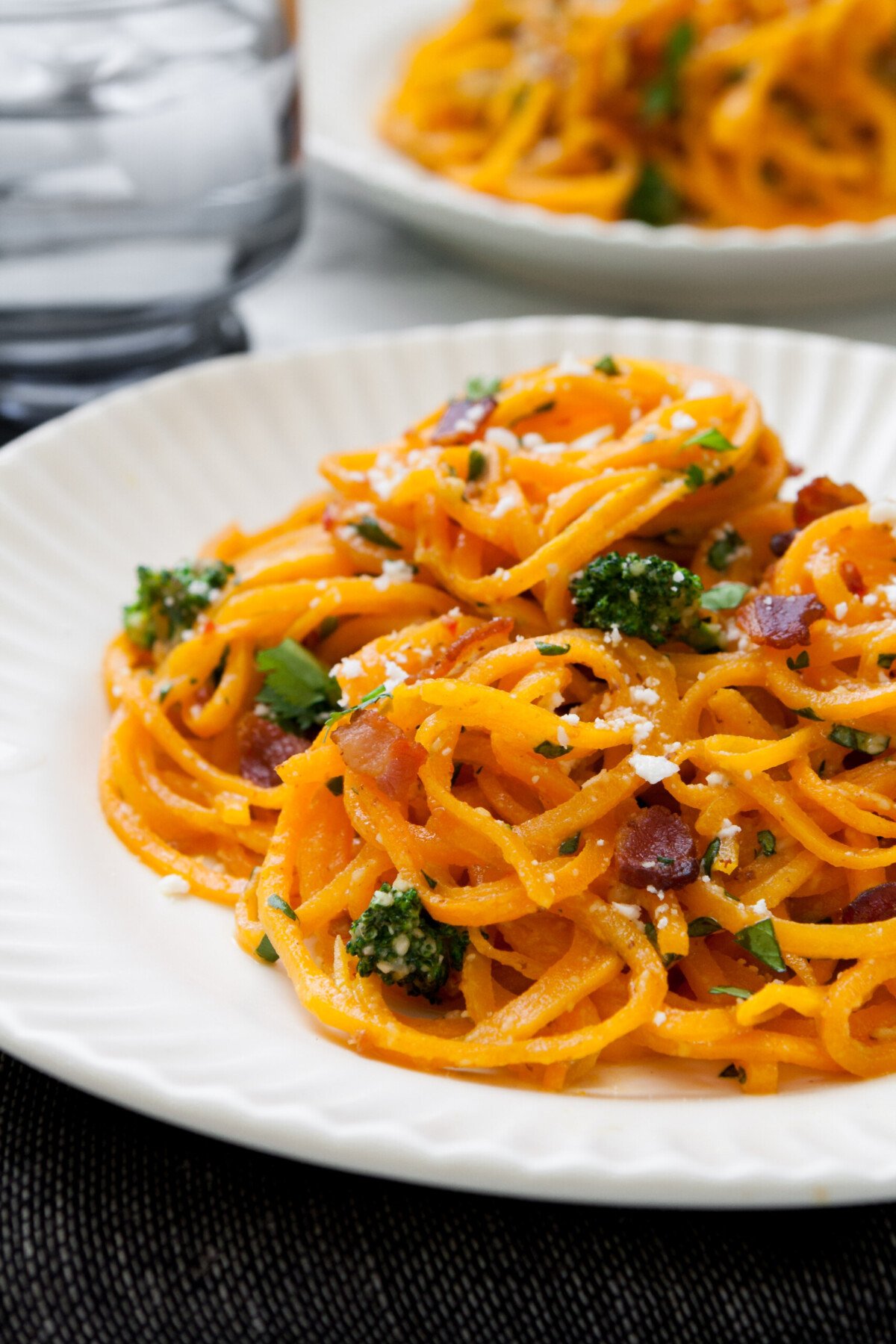 orange noodles coiled on a white plate topped with broccoli and bacon.