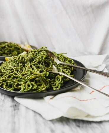 black plate with green pesto-covered pasta on top of marble with tarnished spoon