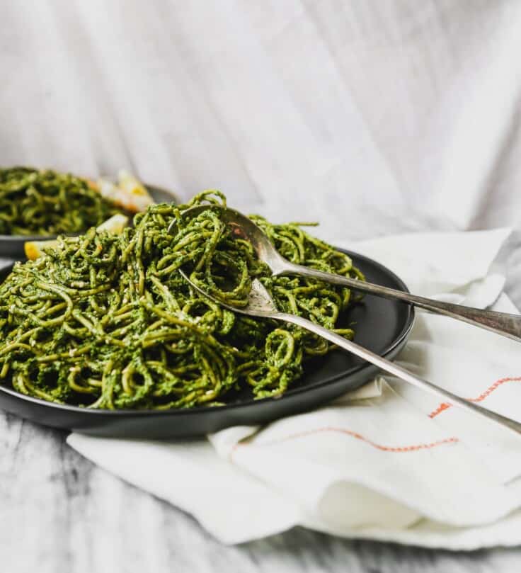 black plate with green pesto-covered pasta on top of marble with tarnished spoon