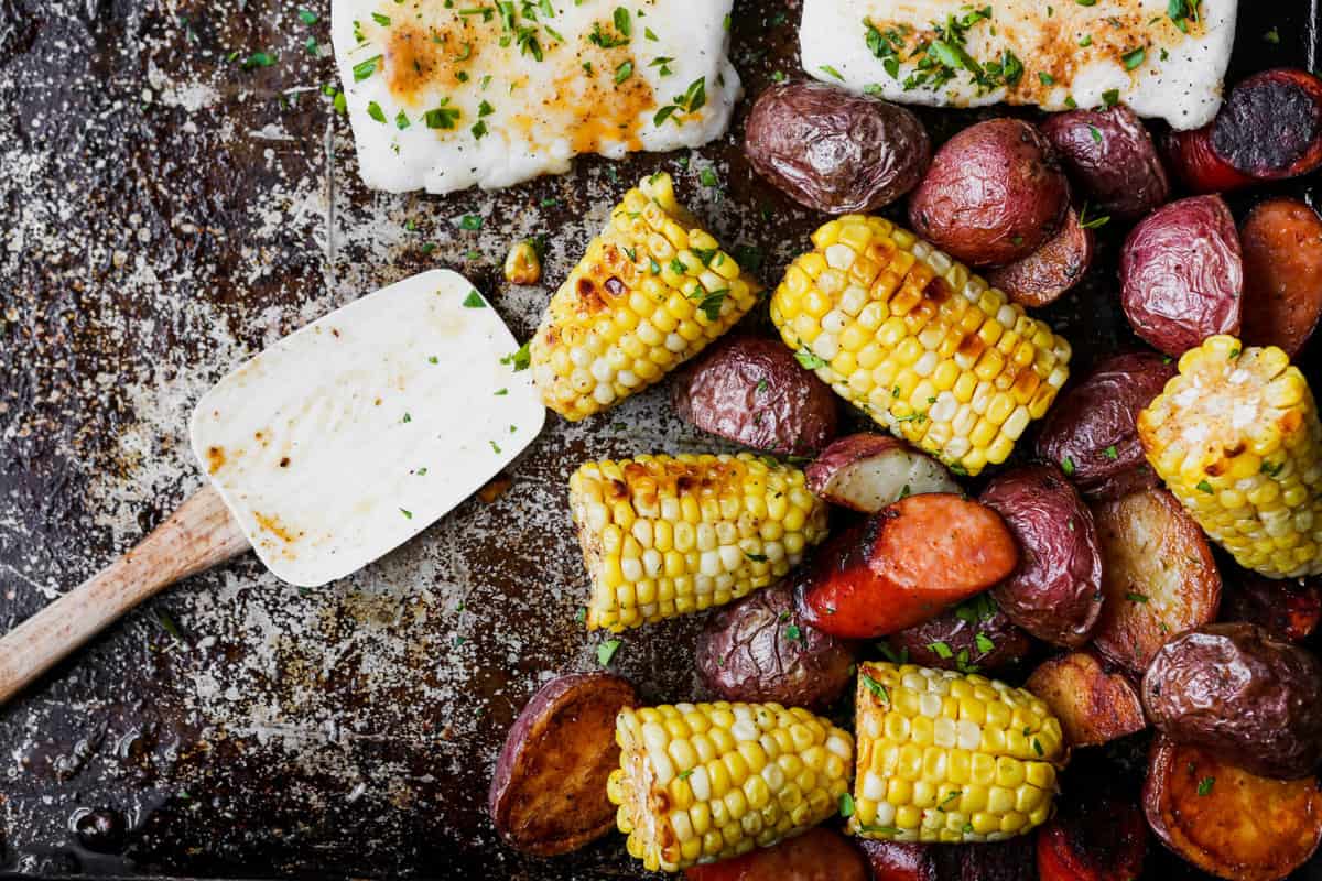 Roasted fish, potatoes, sausage, and corn on worn baking sheet.