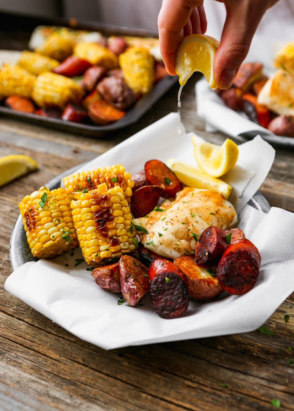 Lemon being squeezed over roasted fish, potatoes, sausage, and corn on paper-lined metal plates set on worn wood surface.