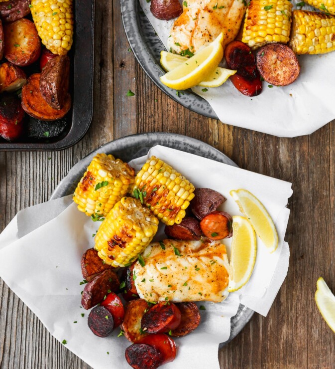 Roasted fish, potatoes, sausage, and corn on paper-lined metal plates set on worn wood surface.
