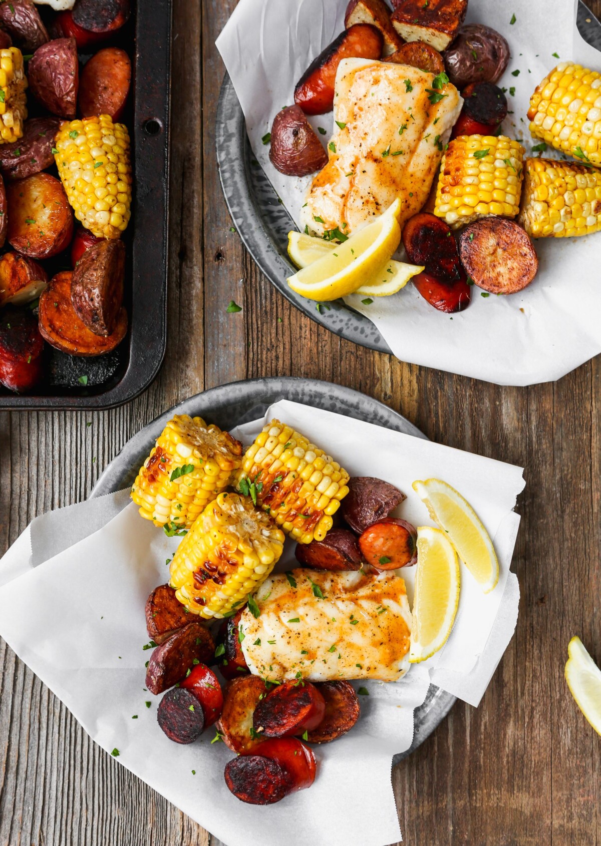 Roasted fish, potatoes, sausage, and corn on paper-lined metal plates set on worn wood surface.