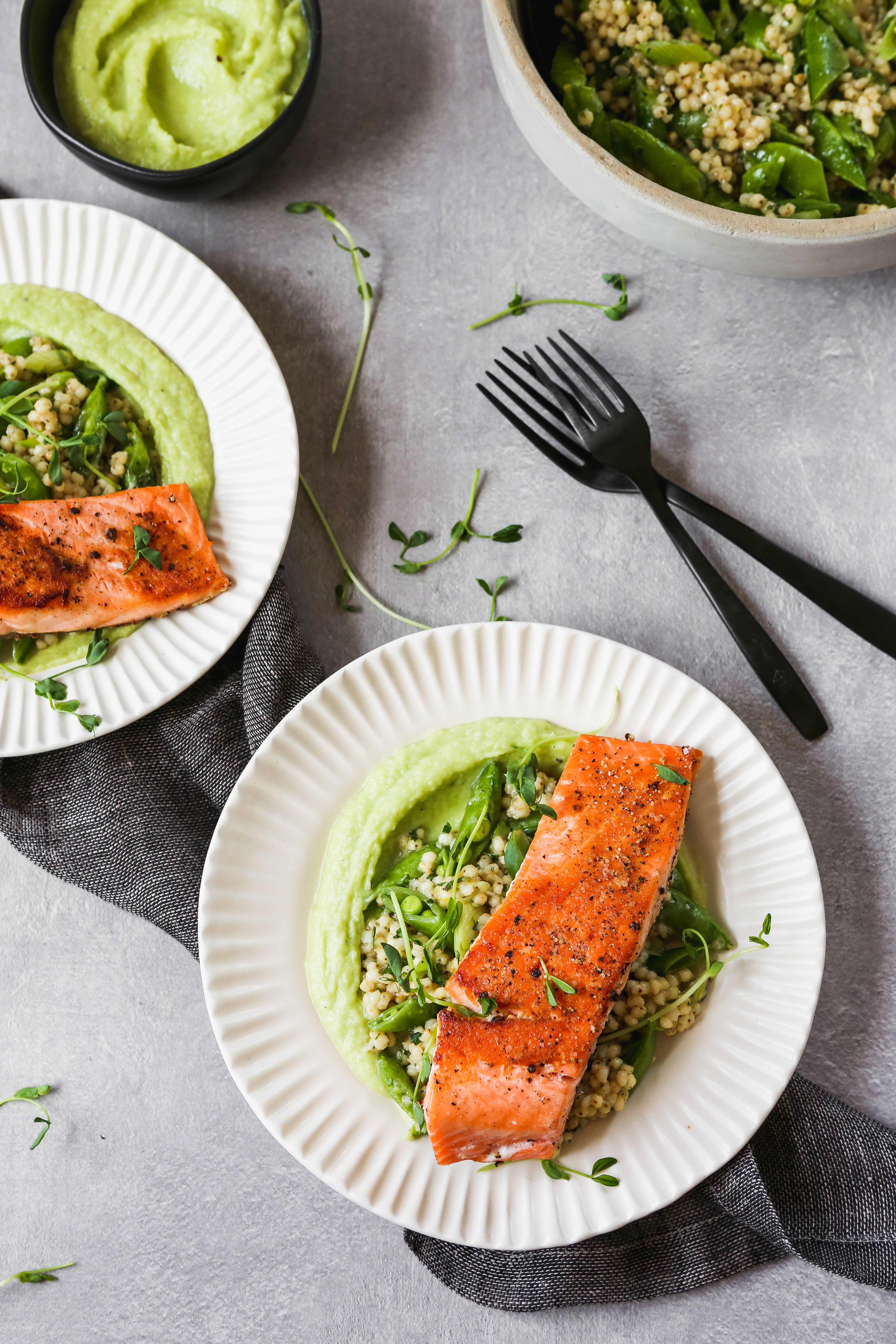 Seared salmon on white plates with green sauce and grain salad, set on a gray background.