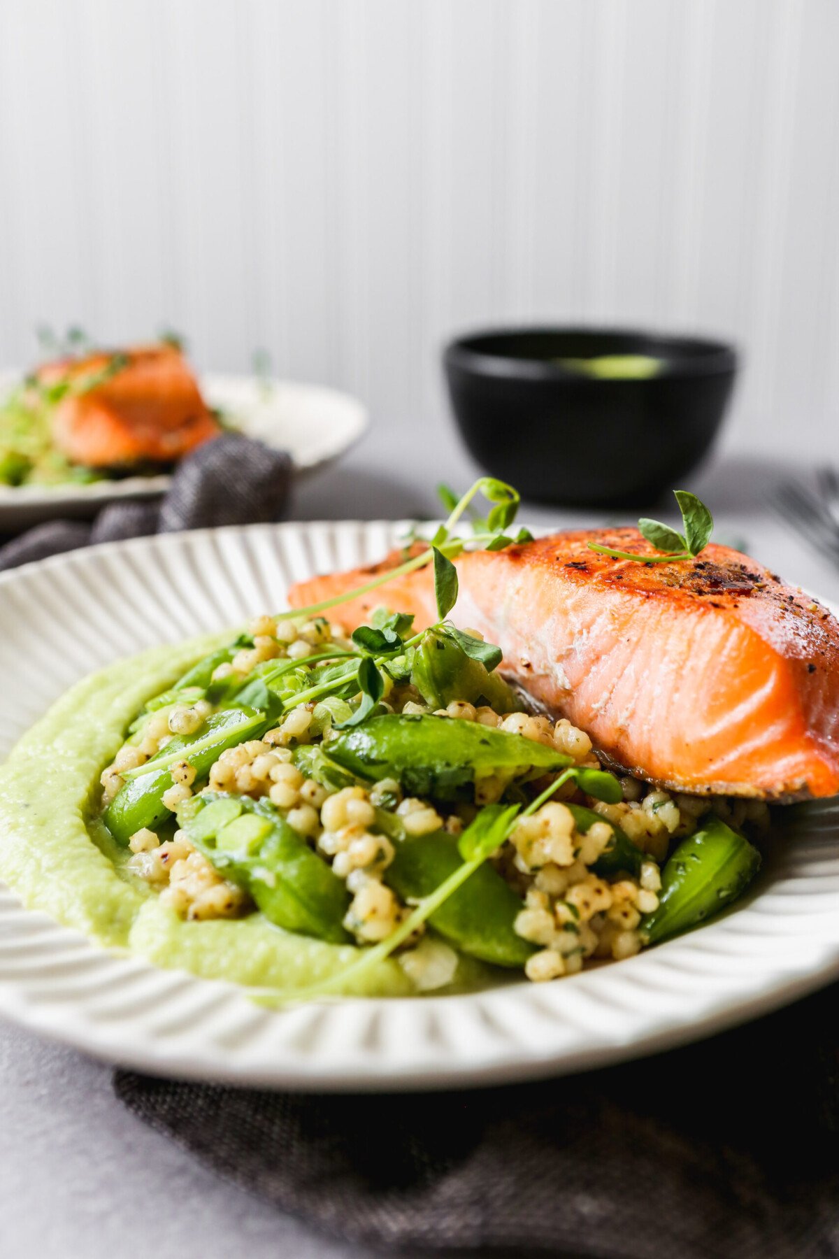 Seared salmon on white plates with green sauce and grain salad, set on a gray background.