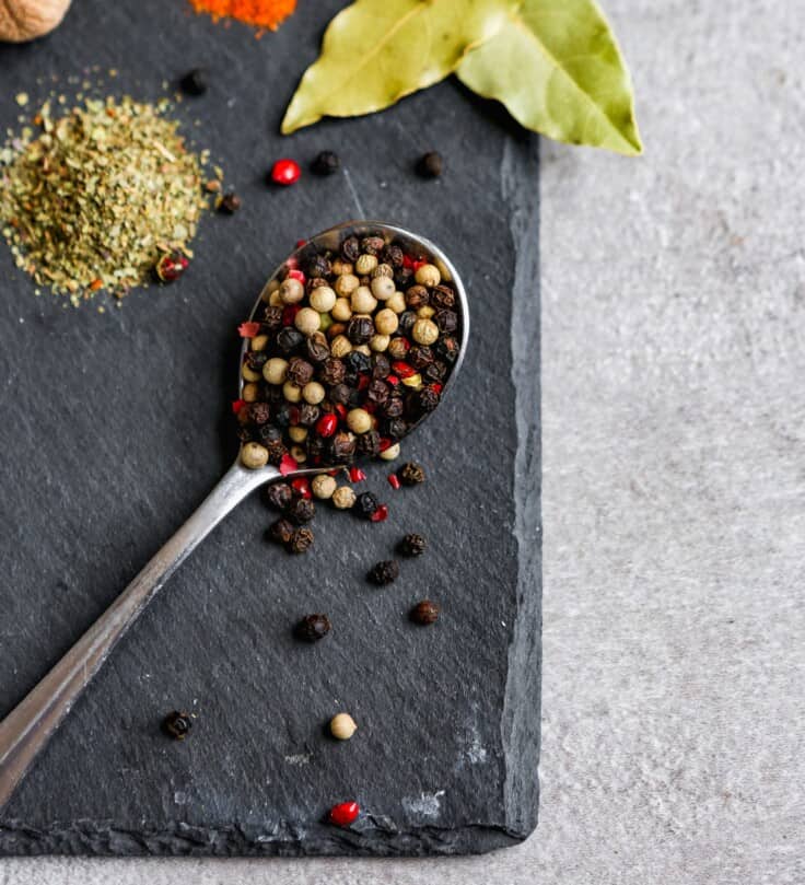 Multi-colored peppercorns spilling off of a spoon on black slate platter
