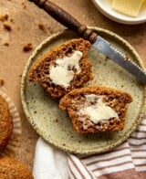 a bran muffin split in half and spread with butter on a speckled plate. A knife set alongside it