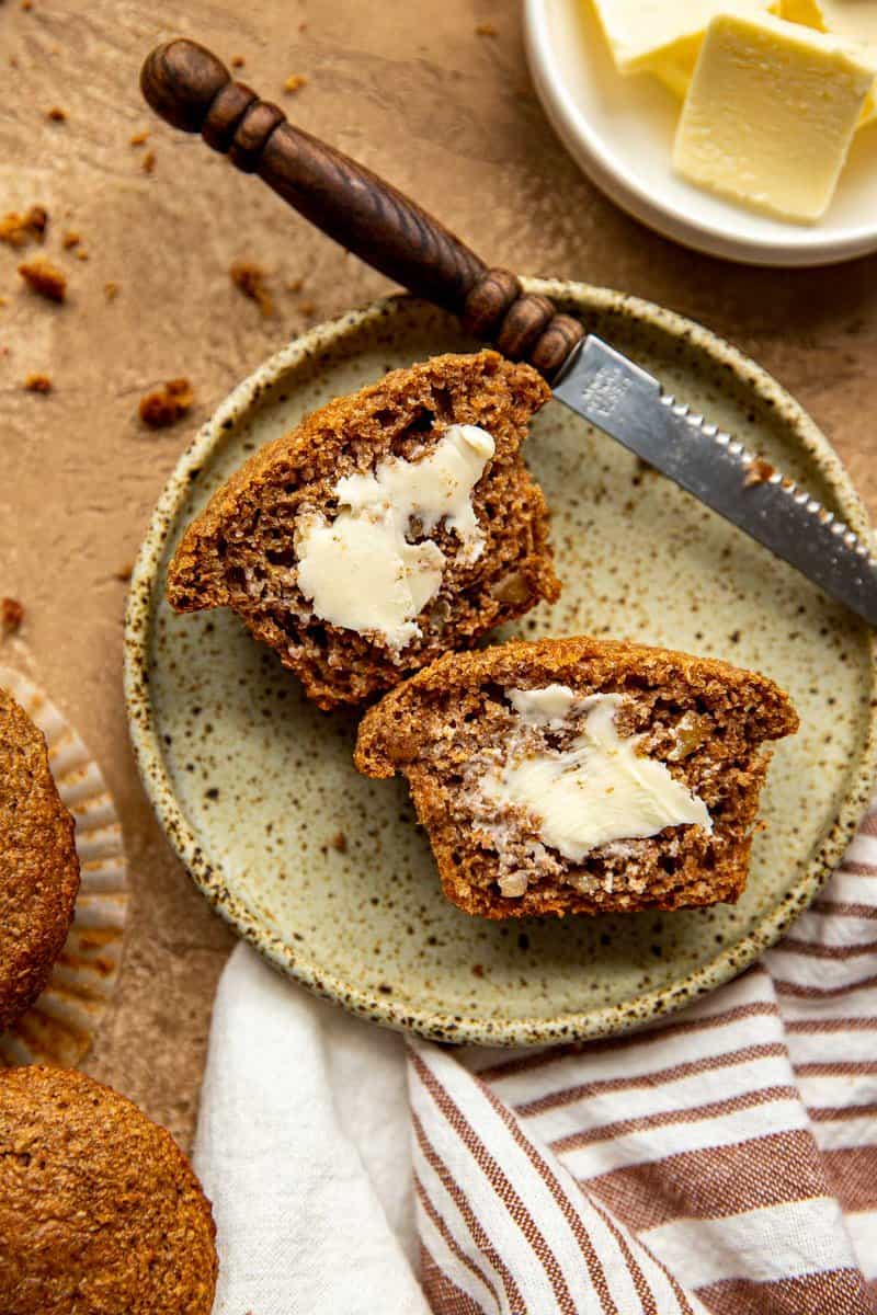 a bran muffin split in half and spread with butter on a speckled plate. A knife set alongside it