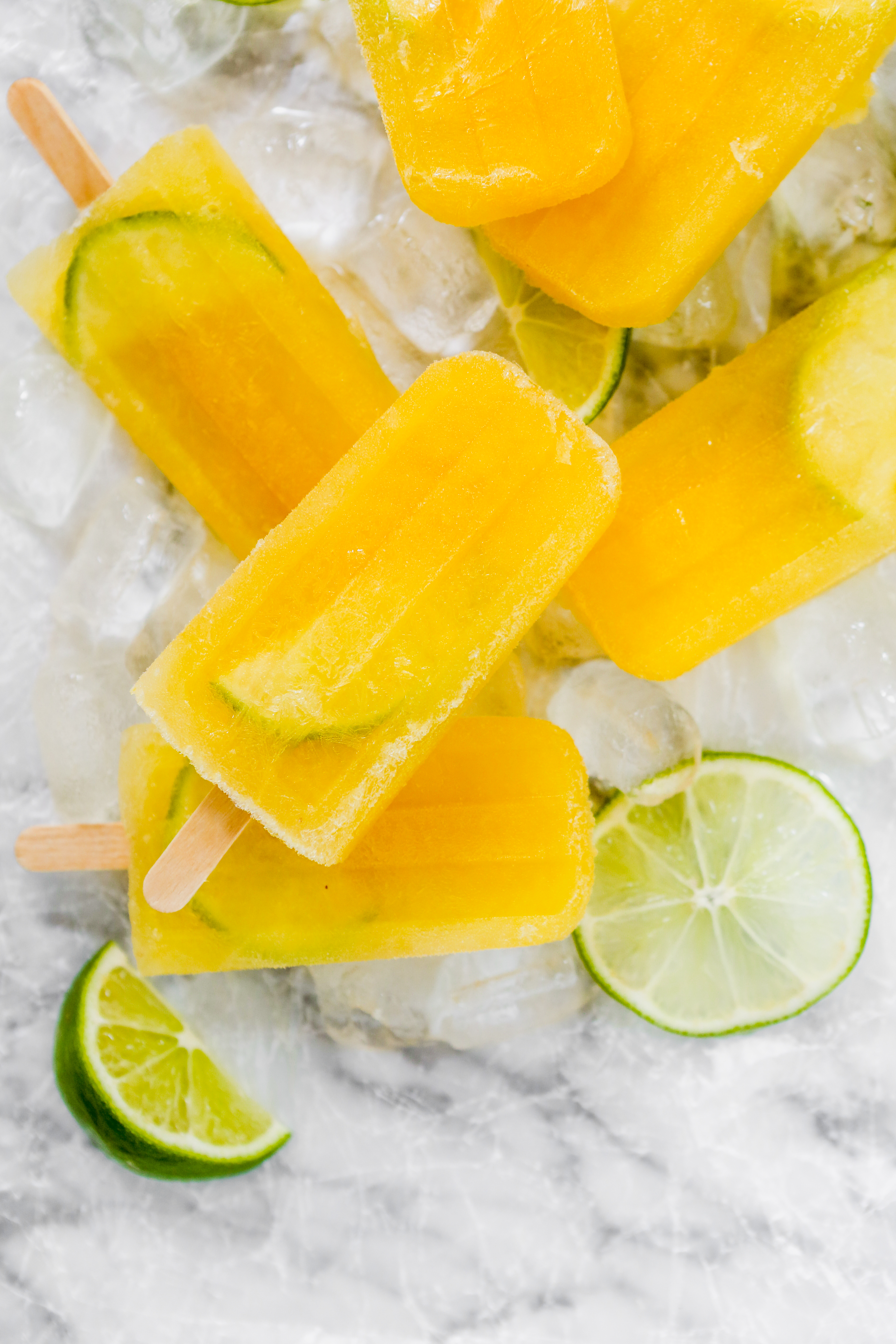Bright yellow popsicles and lime slices set on a white marble surface
