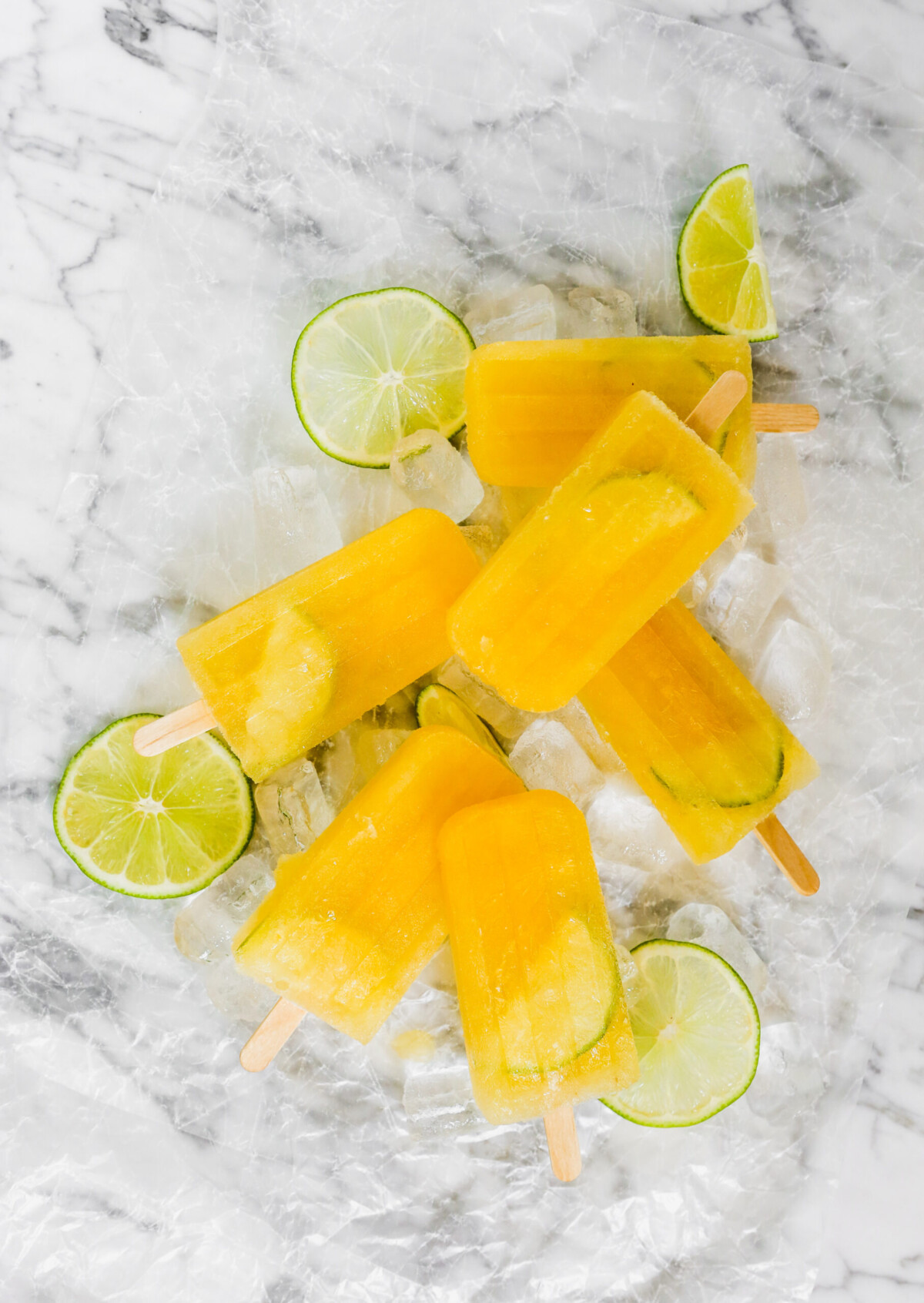 Bright yellow popsicles and lime slices set on a white marble surface