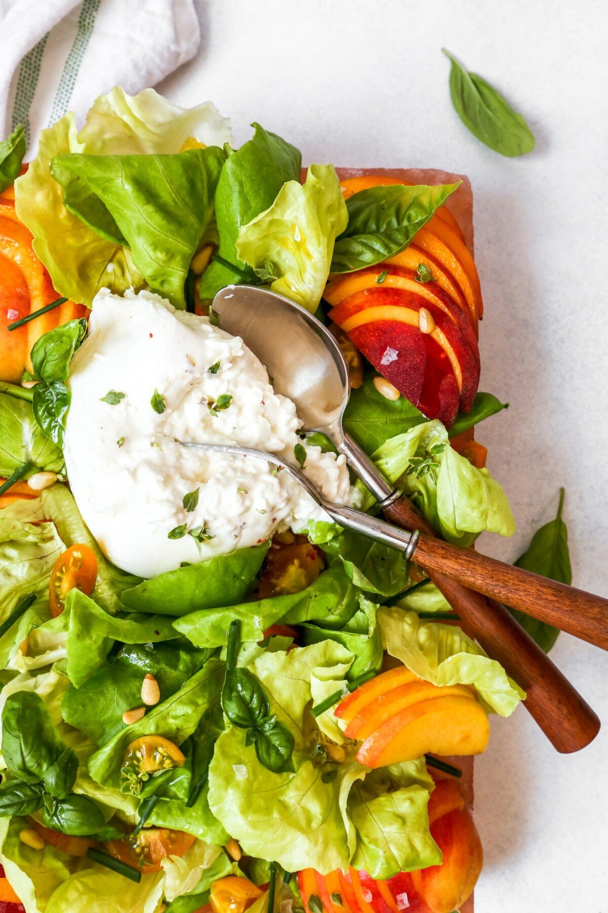 Leafy green salad with peaches, herbs, and burrata arranged on a pink platter, or salt block, set on a white surface.