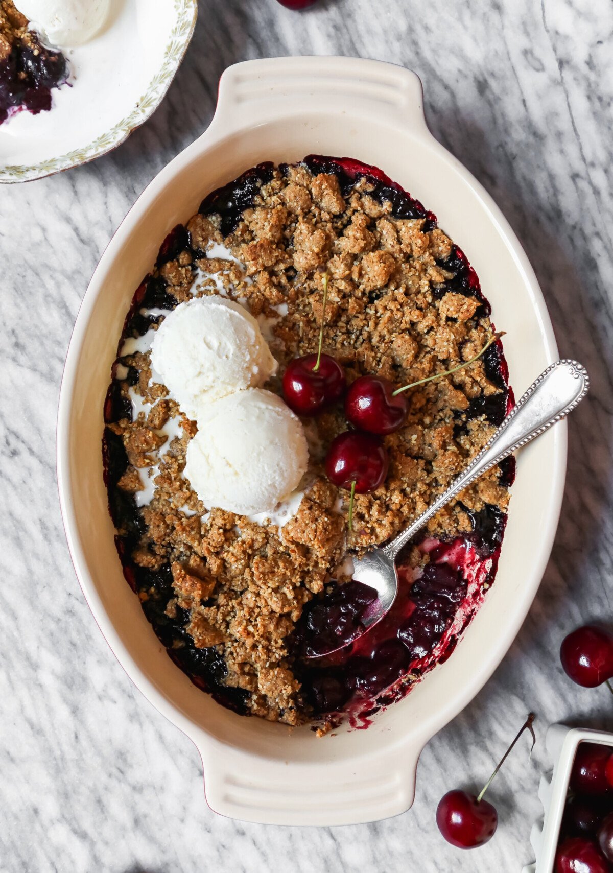 Cherry crisp in an oval baking dish set on a marble surface with servings of cherry crisp and scoops of ice cream in white bowls.