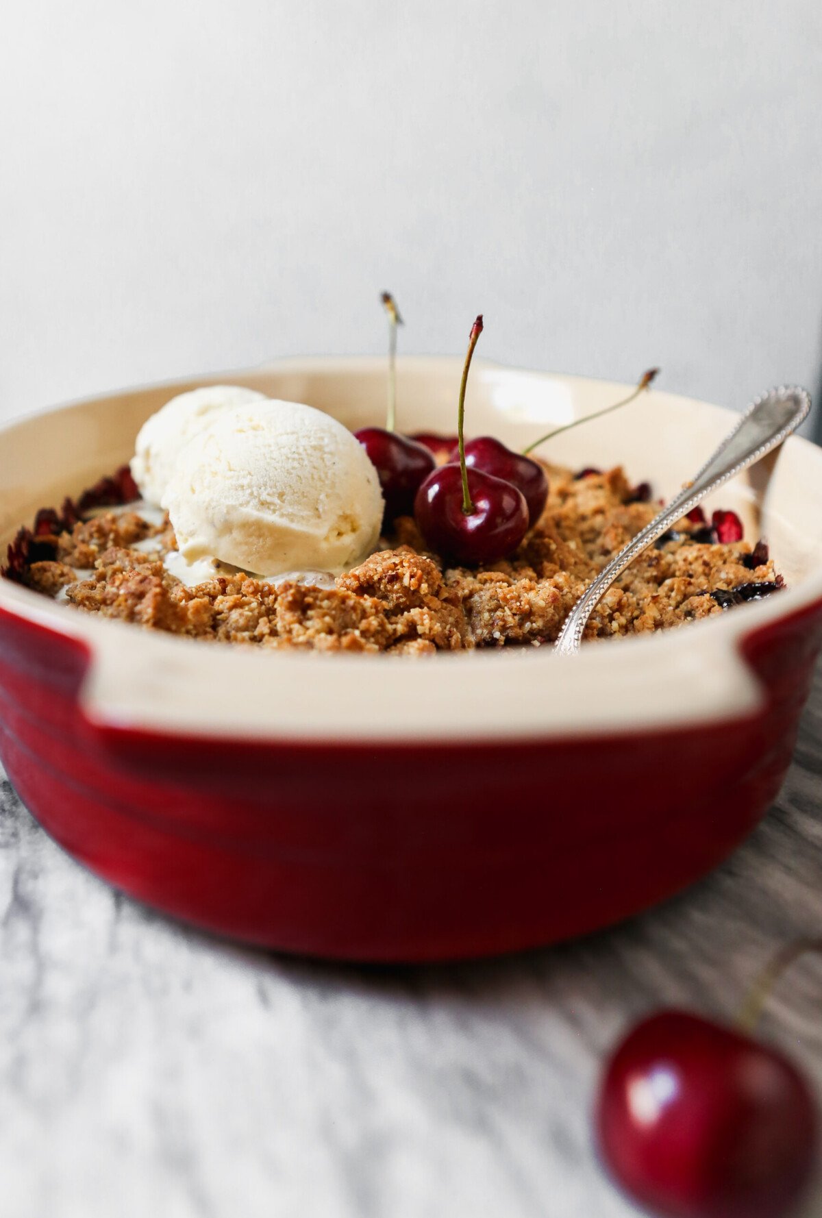 Cherry crisp in an oval baking dish set on a marble surface with servings of cherry crisp and scoops of ice cream in white bowls. 