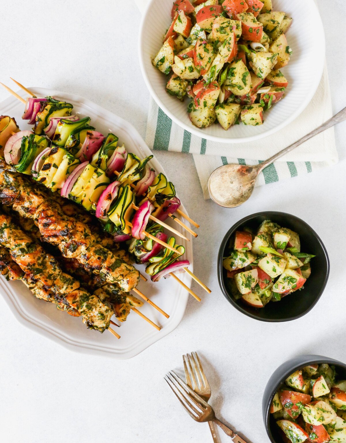 White platter filled with grilled chicken and vegetable skewers, red potato salad, and individual bowls of potato salad on a while background. Summer dinner photography