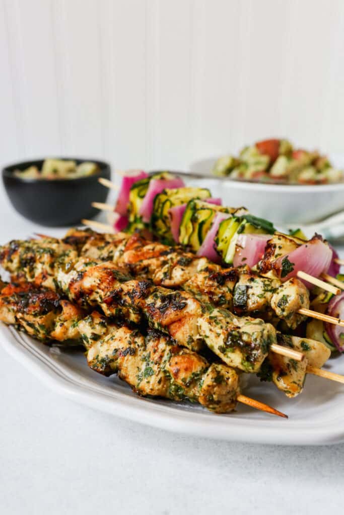 Chicken and vegetable skewers piled on a white platter with a green and white napkin.