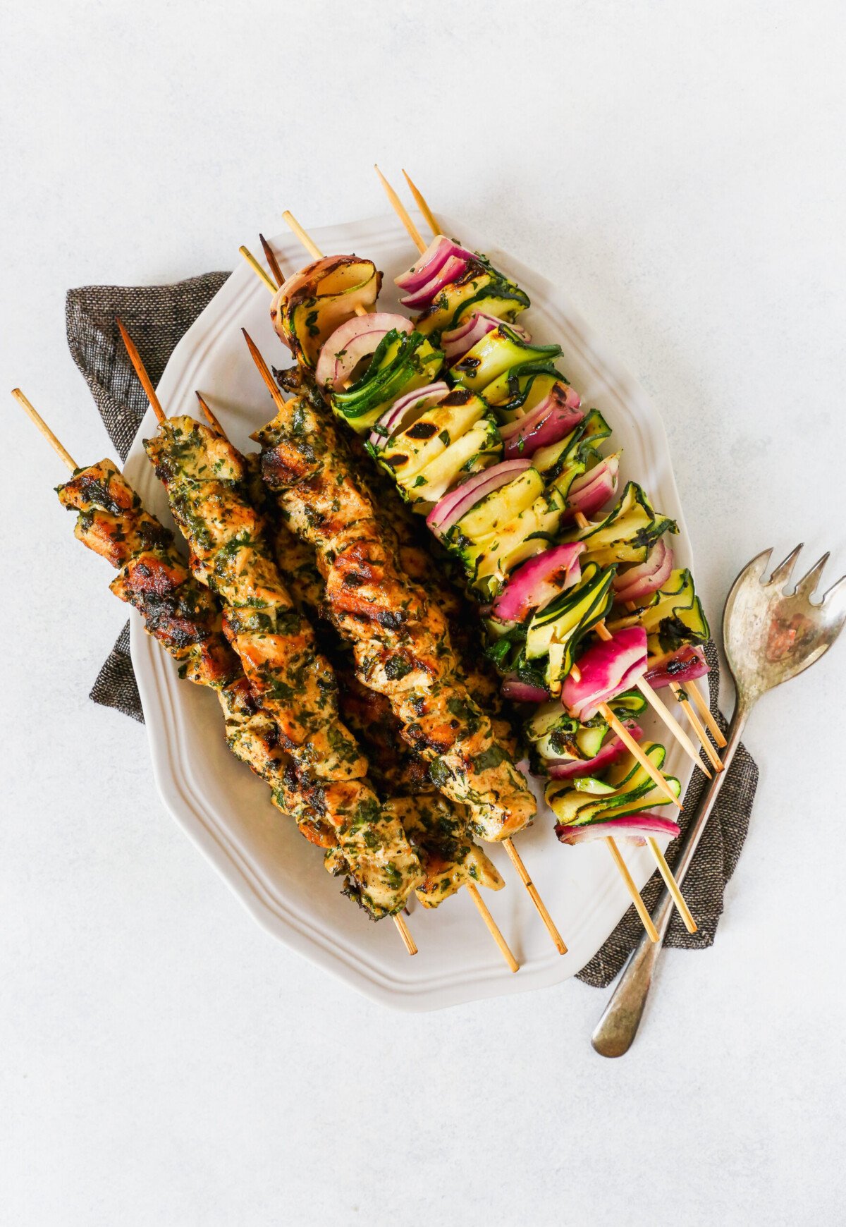 Chicken and vegetable skewers piled on a white platter with a green and white napkin. 
