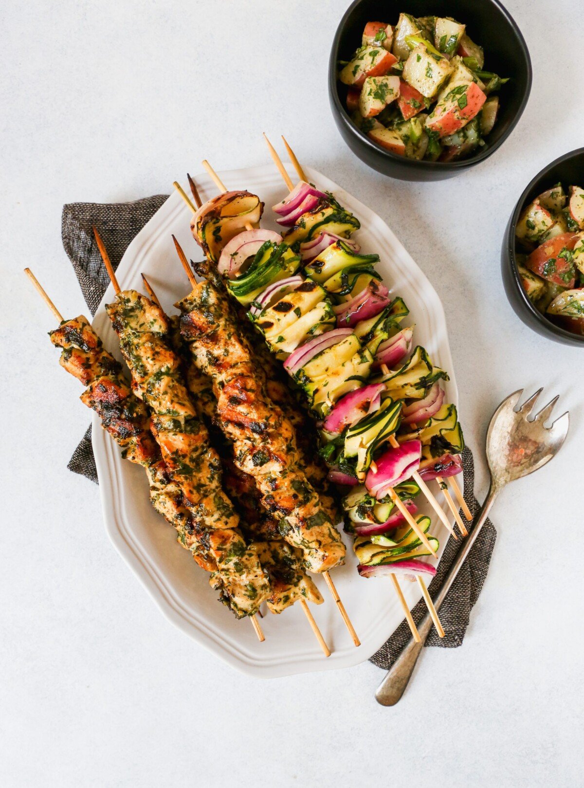 Chicken and vegetable skewers piled on a white platter with a green and white napkin.
