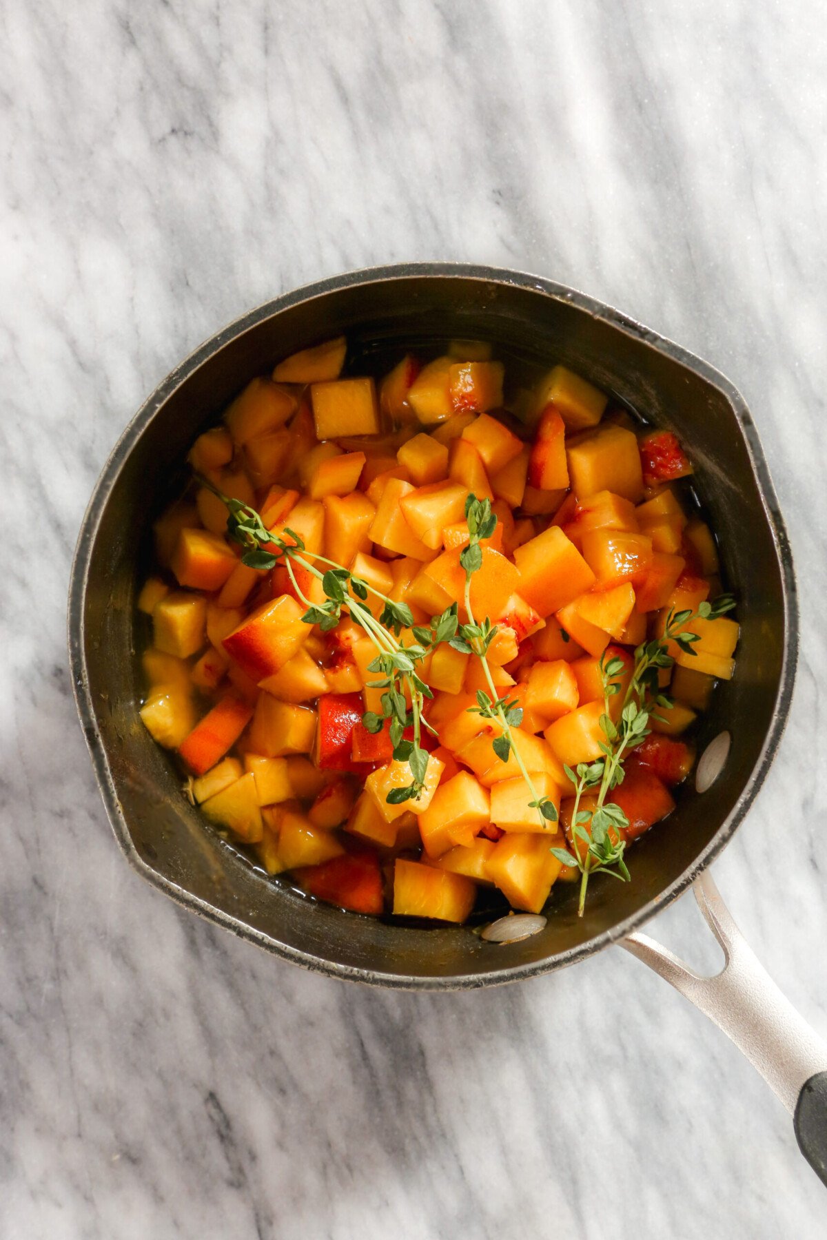 Diced peaches and fresh thyme sprigs in a saucepan set on a marble surface.