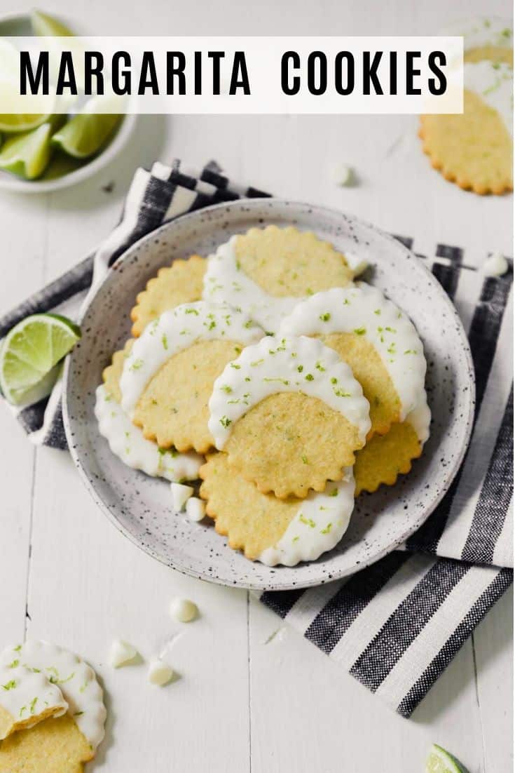 Photograph of glazed cornmeal cookies stacked on a speckled plate on a white table with limes scattered about