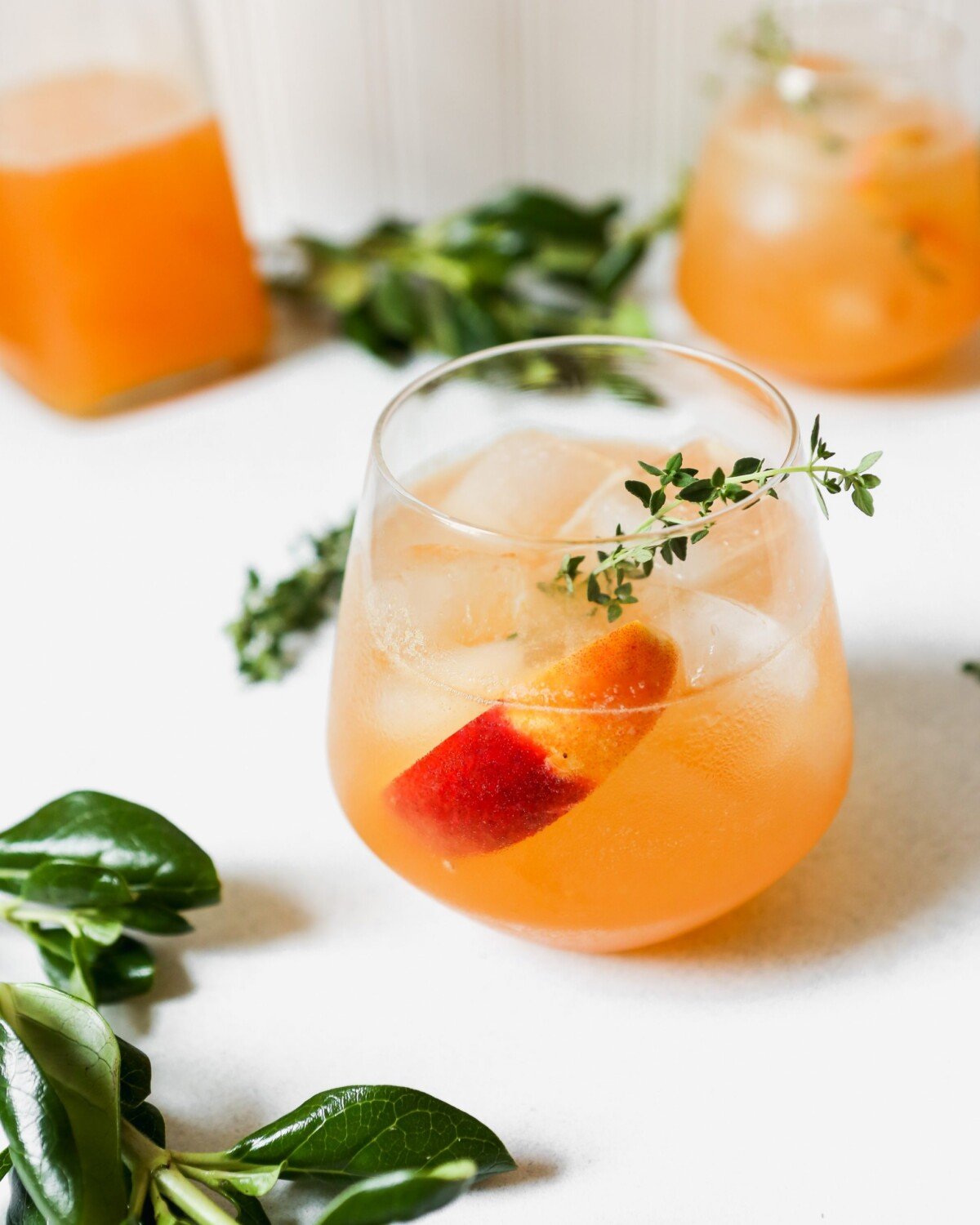 Peach and thyme cocktail photographed from overhead on a white surface. Fresh peach and thyme sprigs arranged around drink.