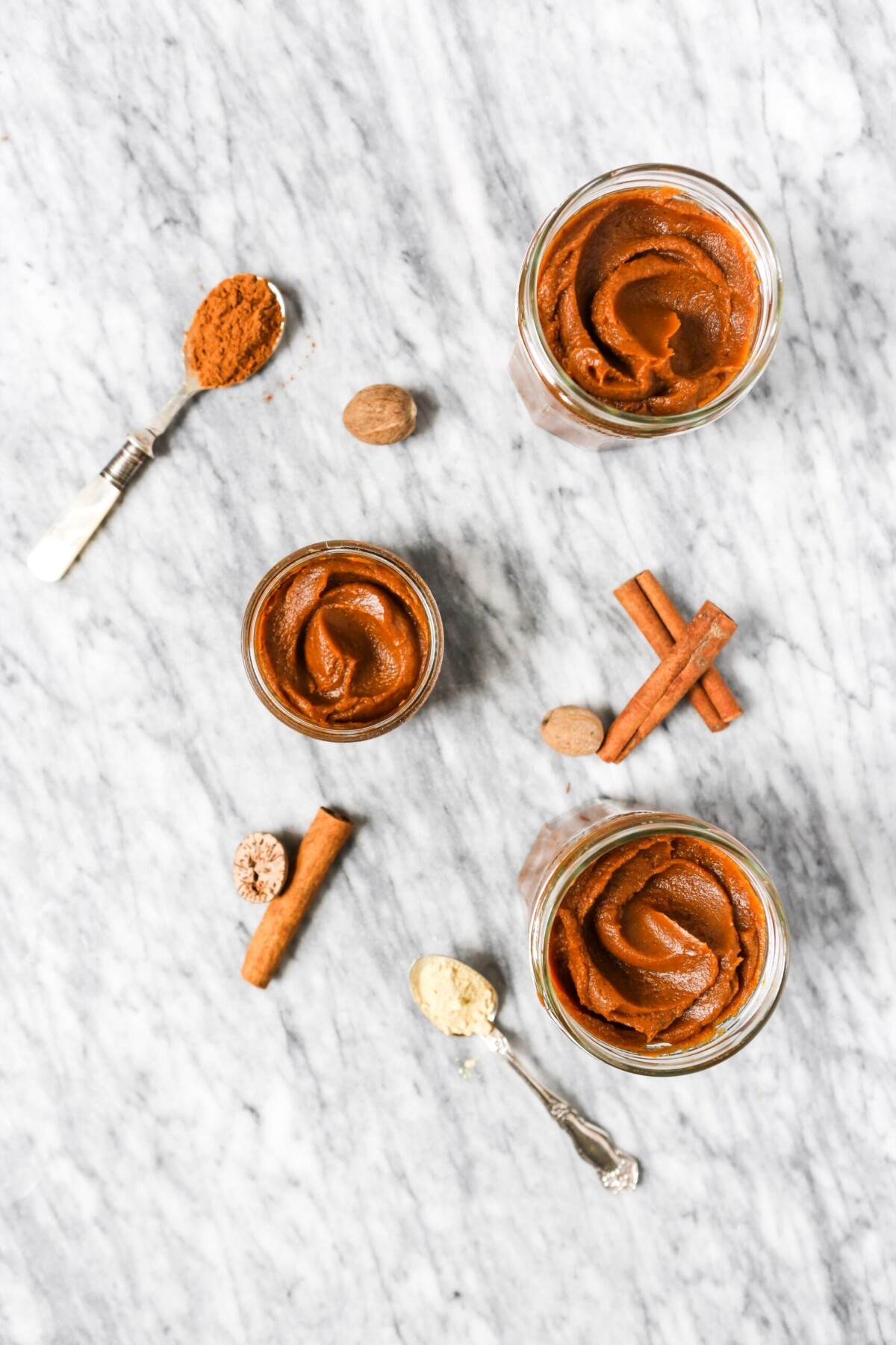 Deep orange pumpkin butter in glass containers on a marble surface with spices scattered around.