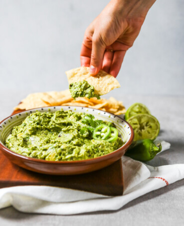 A large bowl of guacamole set on a cutting board with corn chips. Hand dipping chip in guacamole.