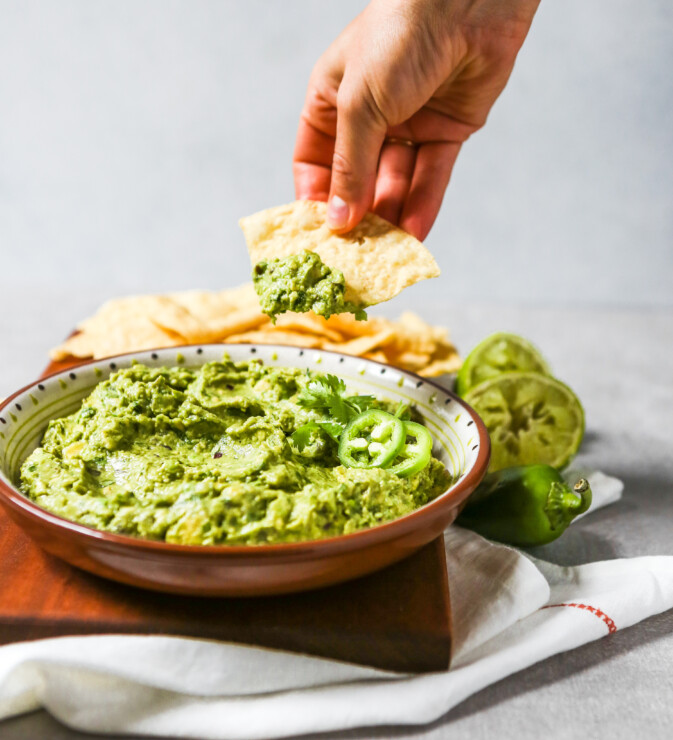 A large bowl of guacamole set on a cutting board with corn chips. Hand dipping chip in guacamole.
