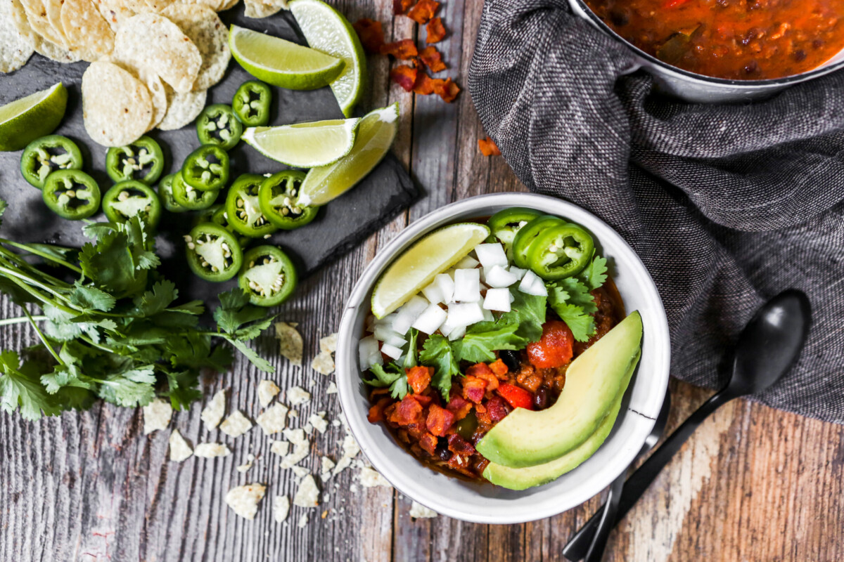 Photo of a bowl of chili topped with avocado, lime, onion, cilantro and jalapeno set on a wood table with chips