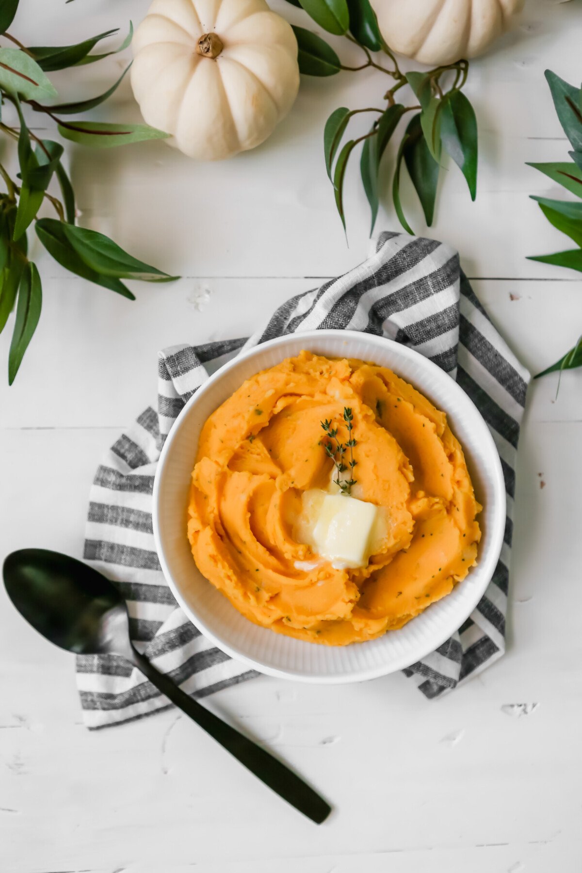 Photograph of a white bowl filled with pumpkin mashed potatoes set on a white table with greenery. 