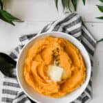 Photograph of a white bowl filled with pumpkin mashed potatoes set on a white table with greenery.