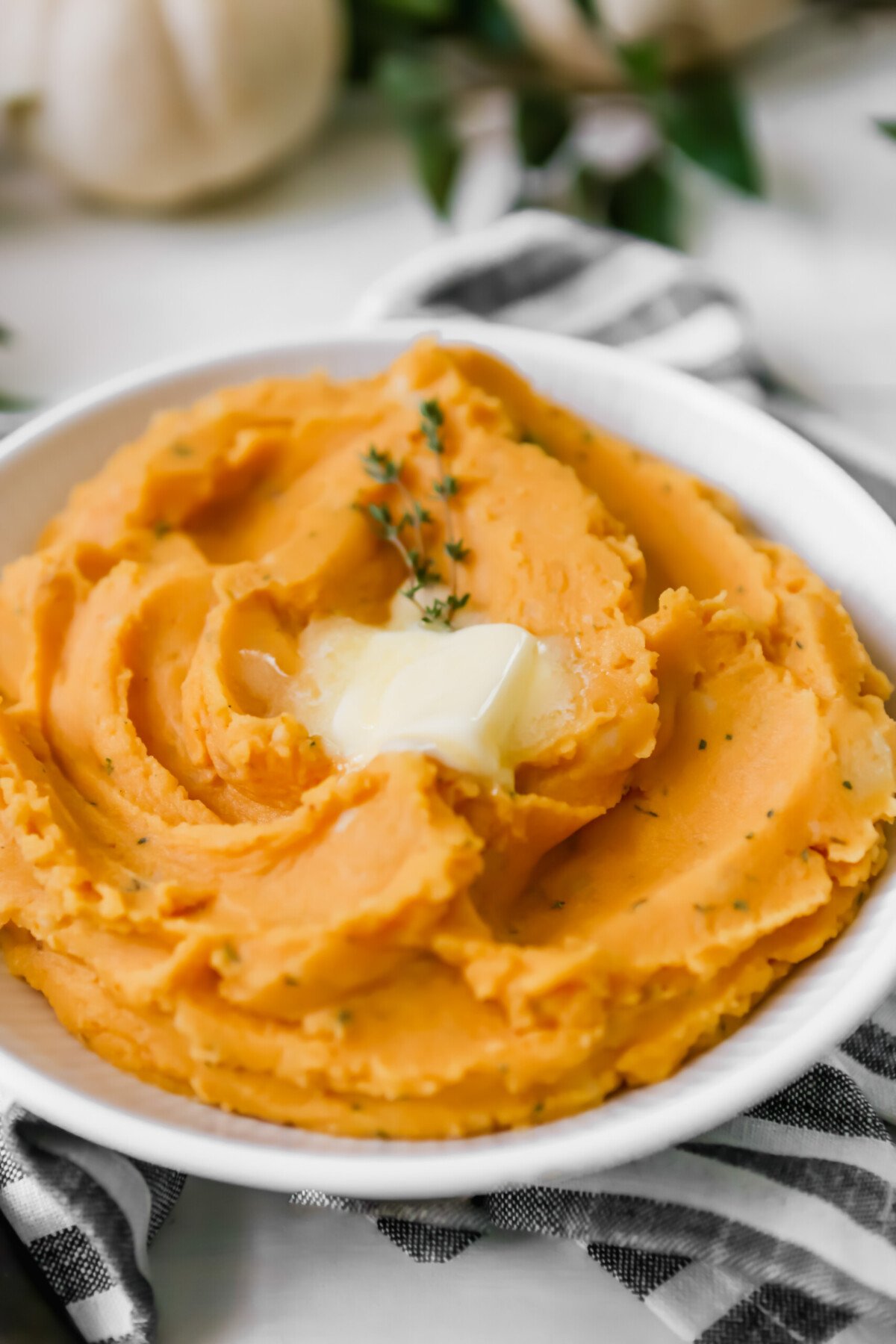Photograph of a white bowl filled with pumpkin mashed potatoes set on a white table with greenery. 