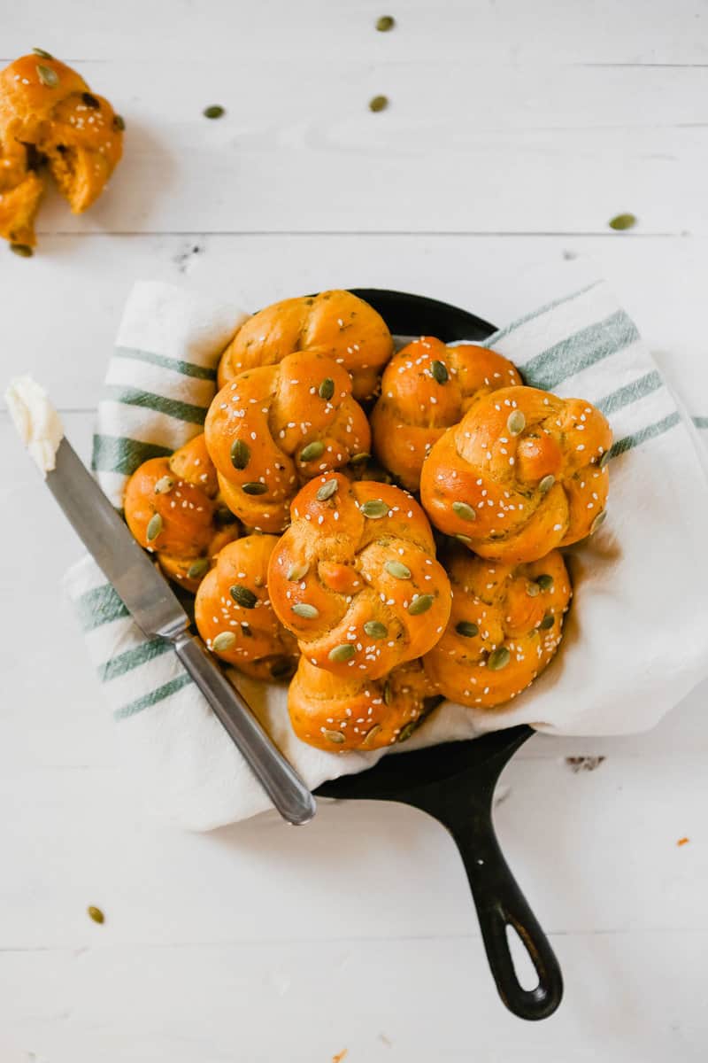 overhead photo of pumpkin challah rolls piled into a cast iron skillet lined with a napkin, a butter knife off to the side